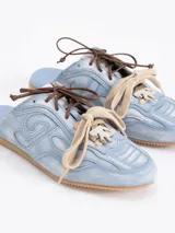 Zapatilla tipo mule (sin talón) de cuero vacuno, color celeste con efecto gastado y detalles de recortes. Presenta un logo estilizado en el lateral y cordones anchos de color beige y marrón.