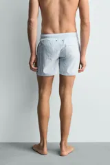 Bañador de hombre de largo medio, color celeste claro con rayas blancas horizontales. Presenta cintura elástica ajustable con cordón, bolsillos frontales y bolsillos traseros de plastrón. Incluye forro interior.