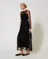 Vestido largo de tul negro con volantes plisados e inserciones de encaje. Presenta un escote redondo con lágrima, manga larga transparente, cremallera invisible lateral, cinturón fino de la misma tela y combinación, de corte ancho.