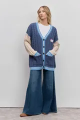 Cardigan de punto con diseño de bloques de color, combinando azul marino en el cuerpo y beige en las mangas. Presenta un tejido con textura de trenzas, cuello en V con borde rayado en azul y blanco, cierre frontal con botones y bolsillos delanteros. Incluye un parche decorativo en el pecho.