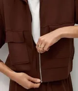 Campera de corte recto y largo, color marrón chocolate, con cuello alto, cierre frontal completo y bolsillos frontales tipo cargo con solapa. Presenta puños y ruedo acanalados.