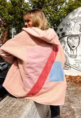 Campera de corderito sintético en color rosa, con diseño de collage en la espalda que incorpora parches de tela en tonos rosa, rojo y azul. Presenta capucha, cierre frontal y un corte holgado y cómodo.