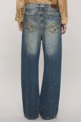 Jeans de tiro medio y calce holgado, color azul con lavado desgastado. Presenta tachas metálicas aplicadas en la parte frontal formando patrones decorativos y en la pretina.