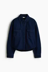 Camisa de denim rígido de algodón azul oscuro, con corte relajado, hombros caídos, cuello clásico, botones ocultos a presión, bolsillos superiores con solapa y bajo redondeado.