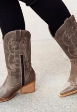 Bota corta de cuero engrasado color marrón con detalle de punta fina y cierre lateral. El interior está forrado en forrería vacuna. El fondo es de madera color negro. Presenta costuras decorativas en color crudo. Tiene un taco de 5 cm.