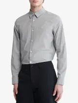 Camisa de vestir gris claro de manga larga, con cuello clásico y cierre de botones. Tiene el logo de Calvin Klein bordado en el pecho.