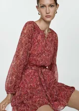 Vestido corto de tejido fluido con estampado floral en tonos rojizos. Presenta cuello redondo con cierre de botones frontal, mangas largas con puños elásticos y lazo ajustable en la cintura. Diseño evasé con forro interior.