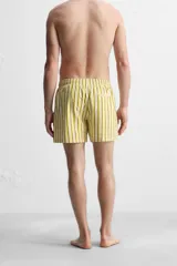 Short de baño confeccionado en tejido técnico, con diseño de rayas verticales amarillas y verdes sobre fondo blanco. Cuenta con cintura elástica ajustable con cordón, bolsillos laterales y bolsillo trasero con vivo.