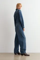 Mono de jean de corte holgado con cierre frontal de botones, cuello camisero y mangas largas. Presenta costuras marcadas y un cinturón de cuero marrón con hebilla metálica en la cintura.