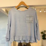 Blusa celeste de manga larga con escote redondo, bolsillo frontal con detalles bordados y volados en el ruedo.