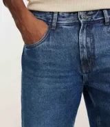 Bermuda de hombre confeccionada en denim de algodón, color azul medio, con tiro medio y corte recto. Presenta cierre de botón y cremallera, bolsillos delanteros y traseros, y etiqueta aplicada en la parte trasera de la cintura.