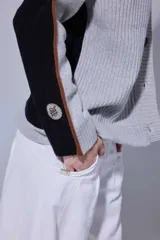 Saco de punto acanalado en color gris claro, con cierre frontal de botones oscuros. Presenta un diseño de bloques de color en las mangas, combinando negro y marrón, y un cuello redondo.