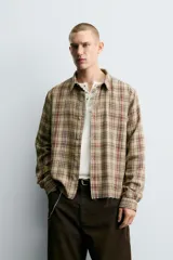 Camisa de corte cropped confeccionada en algodón, con estampado de cuadros en tonos beige, marrón y rojo. Presenta cuello solapa, cierre frontal de botones, manga larga con puño abotonado y detalles de roturas distribuidas por toda la prenda con un bajo irregular.