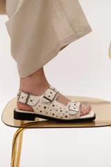 Sandalias negras de cuero con suela plana, dos tiras anchas con hebillas doradas y apliques metálicos.