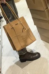 Bolso tipo tote color marrón, confeccionado artesanalmente en cuero vacuno. Presenta un diseño estructurado con costuras decorativas visibles en los bordes y en el centro, y doble asa larga para llevar al hombro.