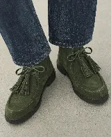 Bota corta de cuero gamuzado en color verde, con diseño de cordones que incluyen borlas decorativas. Presenta una estructura de caña baja, costuras marcadas en la puntera y una suela plana de goma resistente.