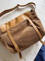 Bandolera tipo messenger bag confeccionada artesanalmente en combinación de cuero y gamuza marrón. Cuenta con solapa frontal de cuero con broches a presión, correas decorativas con hebillas metálicas y una correa larga ajustable para el hombro. Incluye parche con logo grabado en la parte frontal.