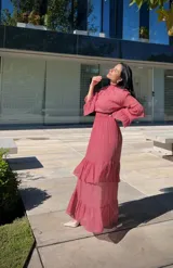 Vestido largo de fiesta color rosa viejo, con cuello alto, mangas largas con volados en los puños y falda con volados escalonados. Presenta una cintura elástica fruncida que define la silueta.