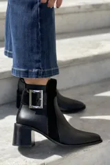 Botín de cuero color topo o gris-marrón, con punta afilada y taco ancho y alto de madera oscura. Presenta un diseño con tira y hebilla metálica cuadrada en el lateral exterior y cierre de cremallera en el lateral interior.