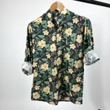 Camisa de hombre con cuello mao, manga larga con puños enrollados y estampado floral en tonos amarillo, verde y violeta sobre fondo negro.