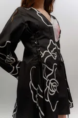 Vestido corto de popelina color negro con estampado de líneas blancas y un diseño floral destacado en el pecho. Cuenta con escote en V, cuello clásico, mangas abombadas y detalles de cordones laterales con efecto corsé.