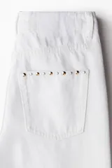 Jean de tiro superalto y corte wide leg, confeccionado en denim de algodón blanco. Presenta diseño de cinco bolsillos, cierre frontal con botón y tachas decorativas distribuidas en patrón por toda la prenda.