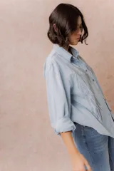 Camisa de denim liviano con calce relajado, cuello camisero clásico y cierre frontal con botones. Presenta una destacada aplicación de strass en el frente formando un diseño de alas.