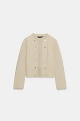 Chaqueta de cuello redondo y manga larga en color beige. Presenta un diseño con doble hilera de botones decorativos en el frente, detalles de encaje a tono y cierre frontal mediante ganchos metálicos ocultos.