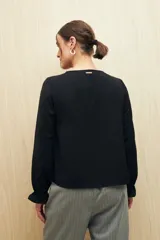 Camisa negra de manga larga con escote en V y botones forrados al tono en el frente. Destaca por su textura sutil y por el detalle de frunces con volados en la parte inferior.