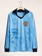 Remera de manga larga en color celeste con estampado de soles y flores de ceibo en tono azul. Presenta cuello polo y puños en tejido rib azul marino, con un parche circular bordado en el pecho que incluye el sol y la inscripción Uruguay Vdamiani.