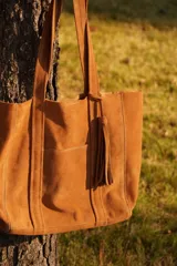 Cartera tote de gamuza color camel, con doble asa de hombro, bolsillo externo y detalle de borla colgante.