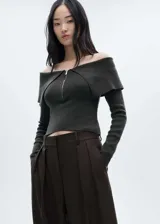 Jersey de punto fino color gris oscuro, con diseño off-shoulder, escote con solapas y cierre frontal de cremallera. Presenta un corte entallado y mangas largas.