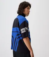 Remera de tricot liviano Blue Steel con corte boxy, cuello polo alto y cierre frontal parcial de un cuarto. Presenta diseño jacquard integral con gráficos y lettering en tonos azul, negro y blanco, mangas cortas y estilo de inspiración deportiva.