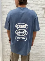 Remera oversize de algodón con efecto acid wash en tono azul oscuro, con estampa blanca en la espalda que incluye los textos "Ost" y "Osten" enmarcados en óvalos, y un diseño de globo terráqueo en el centro.