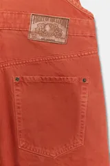Mameluco de denim color naranja con tirantes ajustables, bolsillos de plastrón en el pecho y bolsillos laterales. Cuenta con cierre de ganchos metálicos en el delantero y botones en los laterales.