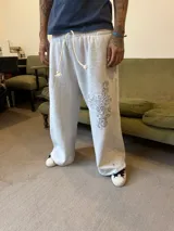 Pantalón de jogging gris de corte baggy, confeccionado en algodón. Presenta cintura elástica con cordón largo ajustable, botones en la parte inferior para ajustar la botamanga y un diseño artístico que combina estampa en plastisol con bordado en relieve sobre el muslo izquierdo.