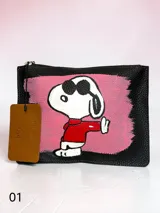 Sobre de cuero negro con textura granulada, intervenido a mano con una ilustración de Snoopy con anteojos y remera roja sobre un fondo pintado en rosa. Incluye etiqueta de cuero colgante con el logo de la colaboración.