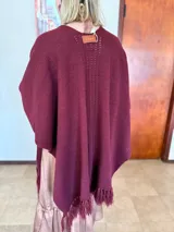 Ruana tejida en color bordó, con diseño de punto acanalado y detalle de calados verticales en la espalda. Presenta terminaciones con flecos en el ruedo y una etiqueta de cuero decorativa en la parte superior.