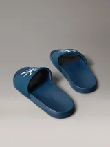 Chinelas de color azul con banda superior de material textil que presenta el logotipo de Calvin Klein bordado en blanco. La suela es ergonómica y antideslizante, con el nombre de la marca grabado en el lateral.