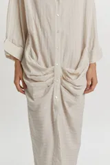 Vestido camisero midi color beige con sutiles rayas verticales oscuras, de manga larga, cuello camisero, cierre frontal con botones y detalle de fruncidos drapeados en los laterales de la falda.
