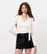 Blusa entallada de microfibra color blanco, con escote en V, mangas largas y detalle de frunces en los laterales. Presenta un lazo integrado en el cuello que permite anudarlo, aportando un toque sofisticado al diseño.