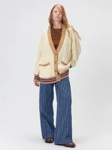 Cardigan de punto oversize en color beige con diseño trenzado. Cuenta con escote en V, cierre frontal de botones, bolsillos delanteros aplicados y detalles en marrón y violeta en la golilla, puños y dobladillo.