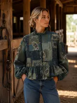 Camisa de lino con estampado patchwork en tonos verdes, azules y tierra. Presenta cuello alto con volado, cierre frontal con botones forrados, mangas largas abullonadas con puños elásticos y un corte evasé con frunce en la cintura.