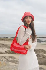 Cartera tipo shoulder bag tejida a crochet en hilo de algodón, con diseño a rayas en tonos de rojo y rosado, y detalles de volados en los bordes y la manija de hombro.