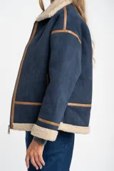 Campera tipo gamulán de corte oversize, color azul marino con interior y cuello de corderito en tono beige. Presenta detalles de costuras y bordes en cuero sintético color marrón, cierre frontal y mangas largas.