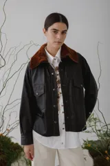 Camisa corta de cuero negro con cuello de piel sintética marrón, cierre frontal con botones a presión, bolsillos con solapa en el pecho y costuras en contraste color naranja.