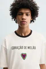 Camiseta de corte relajado confeccionada en algodón, color blanco crudo con cuello redondo en contraste color negro. Presenta un bordado frontal con la frase 'Coração de Melão' y un parche en forma de corazón.