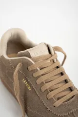 Zapatilla deportiva de gamuza color visón, con cordones a tono y suela de goma marrón. Presenta un diseño retro de running con el nombre de la marca grabado en dorado en el lateral.