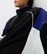 Campera deportiva de poliamida con diseño de bloques de color en negro, blanco y violeta. Presenta cuello alto, cierre frontal con cremallera, puños y ruedo elásticos, y forro interior de poliéster.