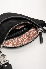 Cartera tipo bandolera de tamaño pequeño, confeccionada en material sintético con textura de gamuza en color marrón topo. Presenta un diseño semicircular con cierre superior, herrajes metálicos plateados y una correa larga ajustable de cinta técnica.